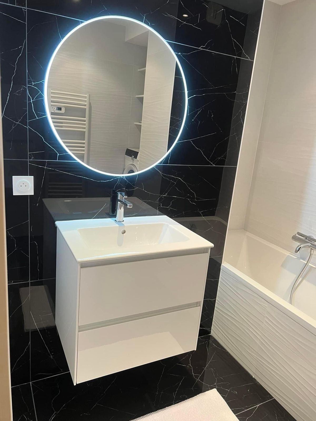 Salle de bain rénovée : grands carreaux effet marbre noir, meuble vasque blanc suspendu, miroir rond lumineux et baignoire habillée de faïence blanche en relief